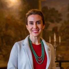 Front-facing of a white woman with brown hair tied back, wearing a red shirt with 3 long, green necklaces and a white blazer, smiling into the camera. Background contains a candelabra and a painting of trees.
