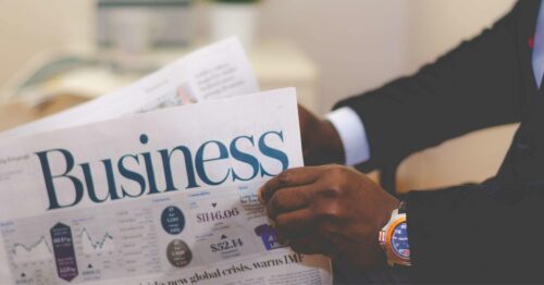 A black person, with a blue and gold watch, holds a copy of the business section of a newspaper