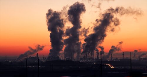 Smoke rises from various industrial plants in a North American city at sunset, bathed in red and orange light